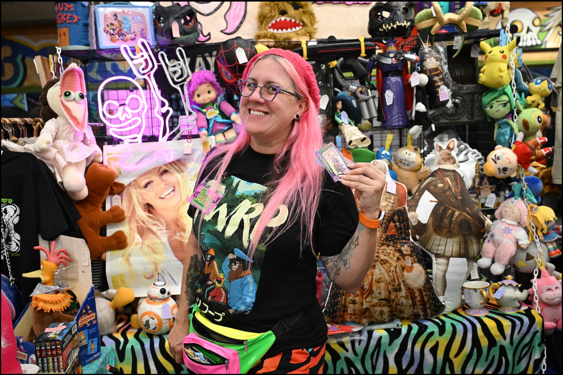 A vendor with pink hair smiling behind a booth packed with toys and collectibles
